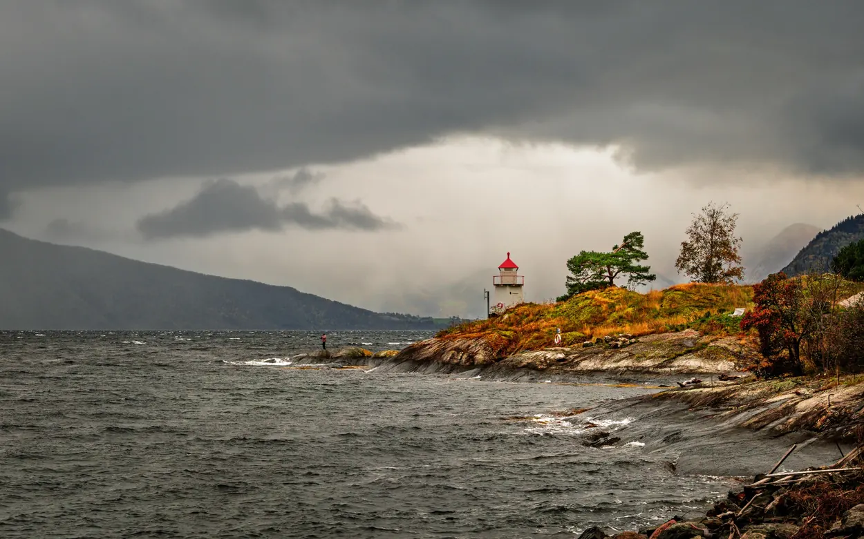 Small white lighthouse with a red roof on a rocky Norwegian coastline beneath heavy rain clouds