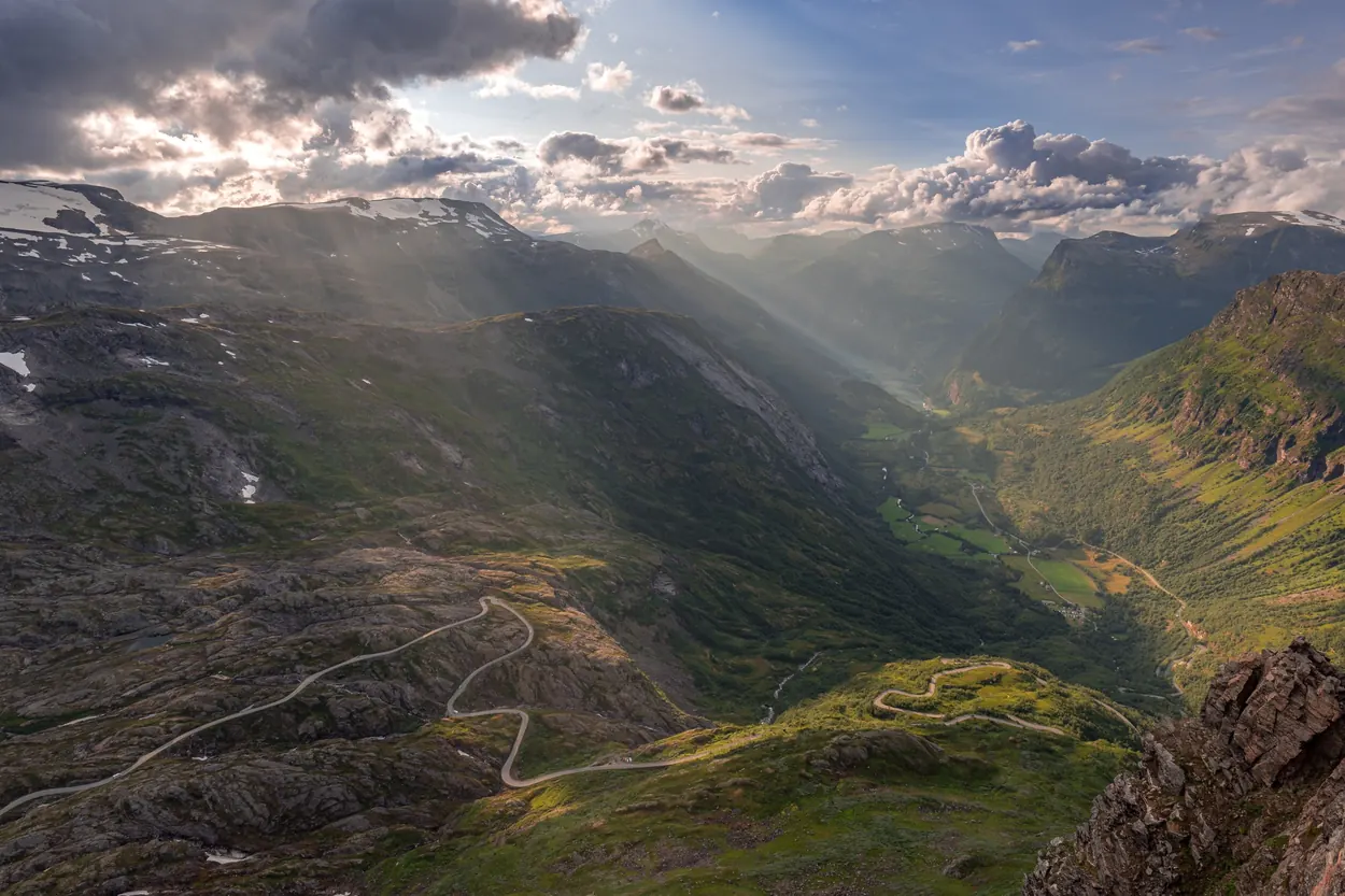 Winding mountain road descending into a deep green valley beneath dramatic clouds